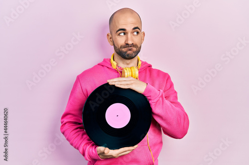 Young bald man holding vinyl disc smiling looking to the side and staring away thinking.