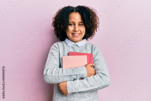 Young little girl with afro hair holding books winking looking at the camera with sexy expression, cheerful and happy face.