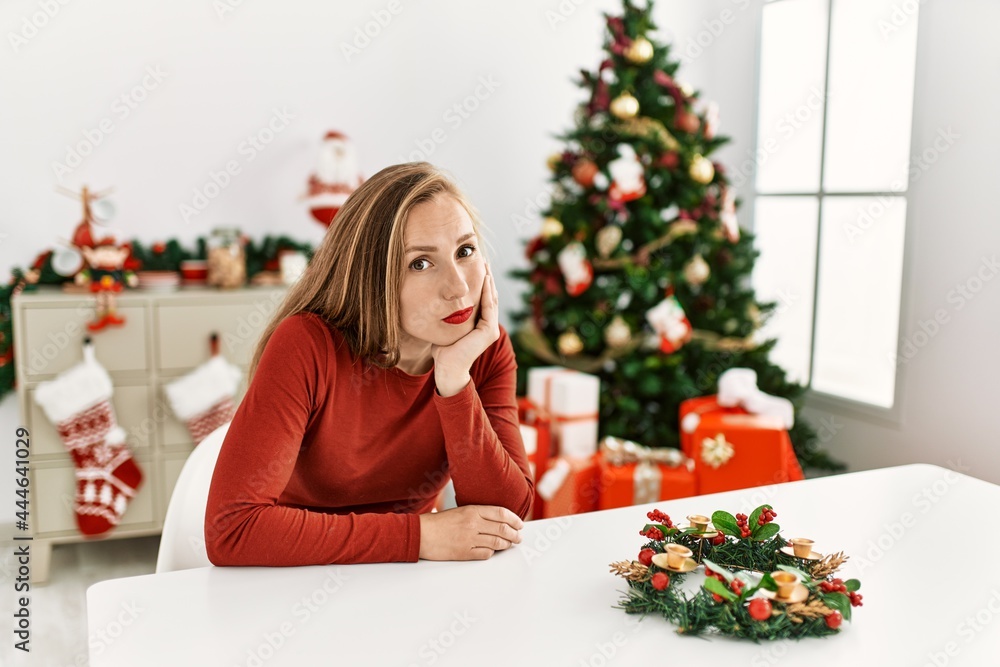 Caucasian young blonde woman sitting on the table by christmas tree thinking looking tired and bored with depression problems with crossed arms. Caucasian young blonde woman sitting on the table by christmas tree thinking looking tired and bored with depression problems with crossed arms.