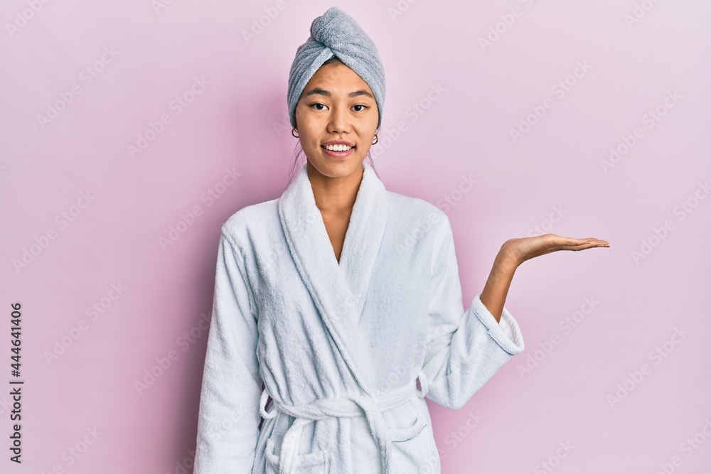 Young chinese woman wearing shower towel cap and bathrobe smiling cheerful presenting and pointing with palm of hand looking at the camera. Young chinese woman wearing shower towel cap and bathrobe smiling cheerful presenting and pointing with palm of hand looking at the camera.