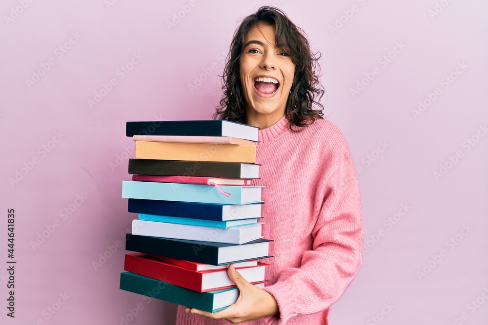 Young hispanic woman holding a pile of books celebrating crazy and amazed for success with open eyes screaming excited. Young hispanic woman holding a pile of books celebrating crazy and amazed for success with open eyes screaming excited.