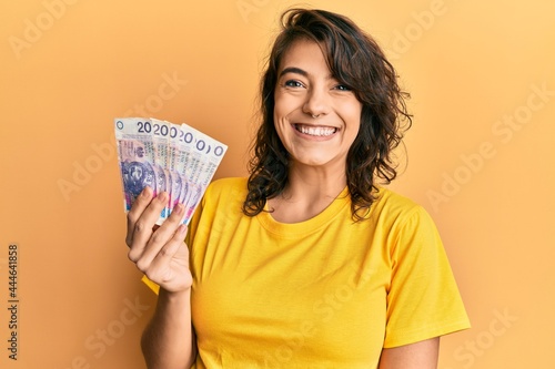 Young hispanic woman holding 20 polish zloty banknotes looking positive and happy standing and smiling with a confident smile showing teeth