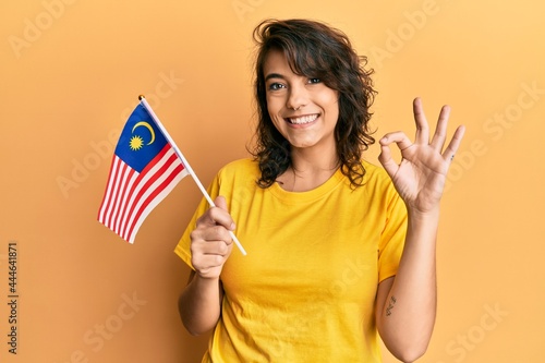 Young hispanic woman holding malaysia flag doing ok sign with fingers  smiling friendly gesturing excellent symbol