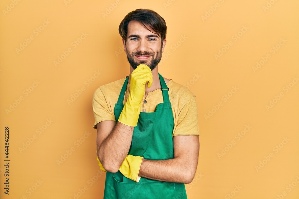 Young hispanic man wearing cleaner apron and gloves smiling looking confident at the camera with crossed arms and hand on chin. thinking positive. Young hispanic man wearing cleaner apron and gloves smiling looking confident at the camera with crossed arms and hand on chin. thinking positive.