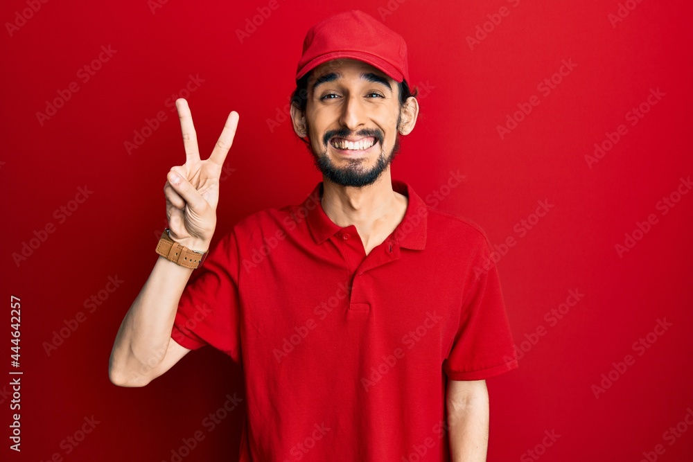 Young hispanic man wearing delivery uniform and cap smiling with happy face winking at the camera doing victory sign. number two.