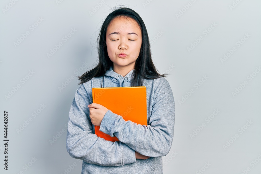 Young chinese girl holding book looking at the camera blowing a kiss being lovely and sexy. love expression. Young chinese girl holding book looking at the camera blowing a kiss being lovely and sexy. love expression.
