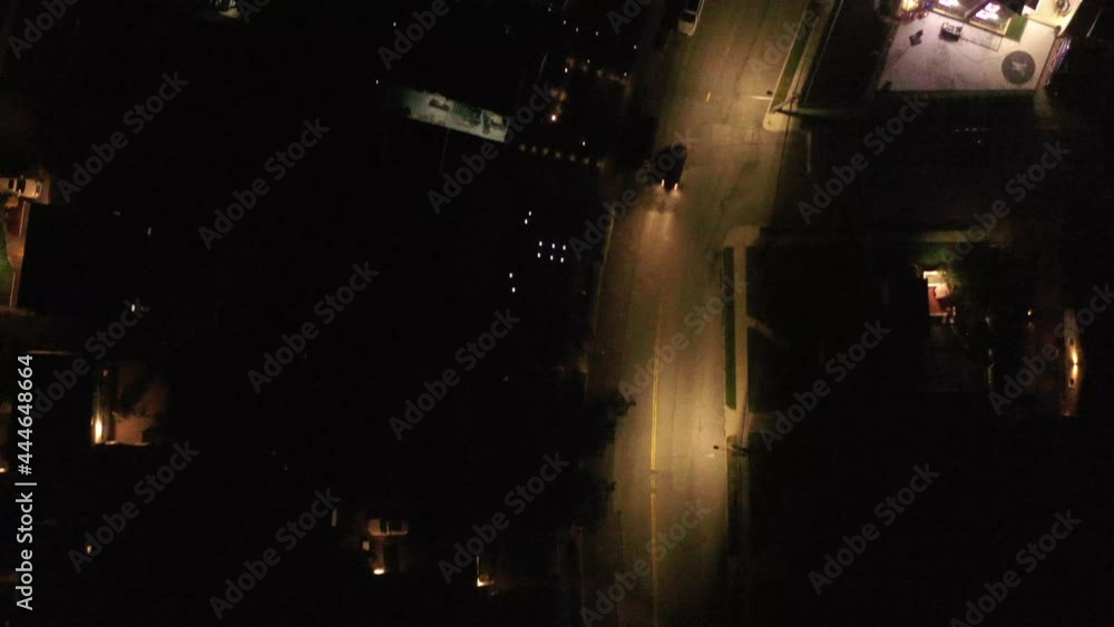 A top down shot directly above a two lane street, illuminated by streetlights in a quiet suburban neighborhood. A car drives by below as the drone camera tilted down dolly in. It moves forward slowly. A top down shot directly above a two lane street, illuminated by streetlights in a quiet suburban neighborhood. A car drives by below as the drone camera tilted down dolly in. It moves forward slowly.