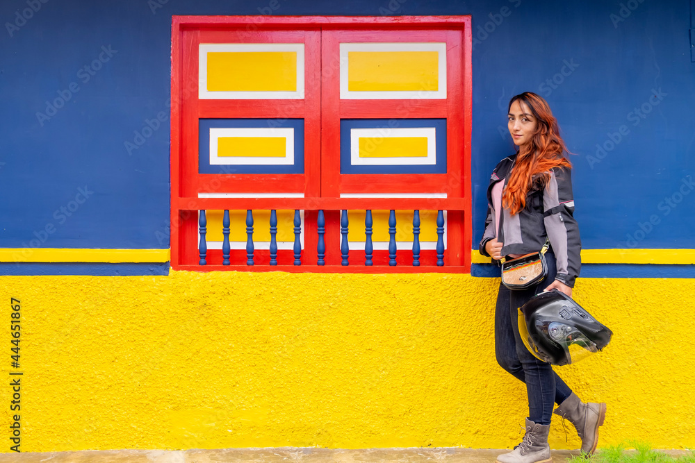 red-haired woman, with a motorcycle helmet in hand, next to a red, yellow and blue window