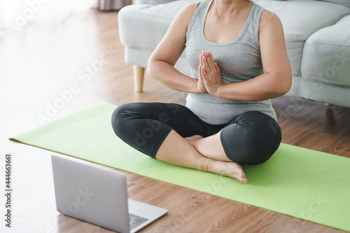 Fototapeta Naklejka Na Ścianę i Meble -  Mature Asian chubby fat woman sitting on the floor in living room practice online yoga lesson with the computer. female having meditate training class on the laptop..