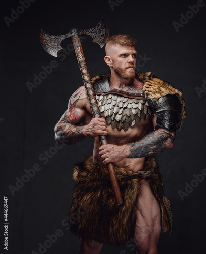 Brutal tattooed warrior wearing light armour and fur holding two-handed axe in dark studio