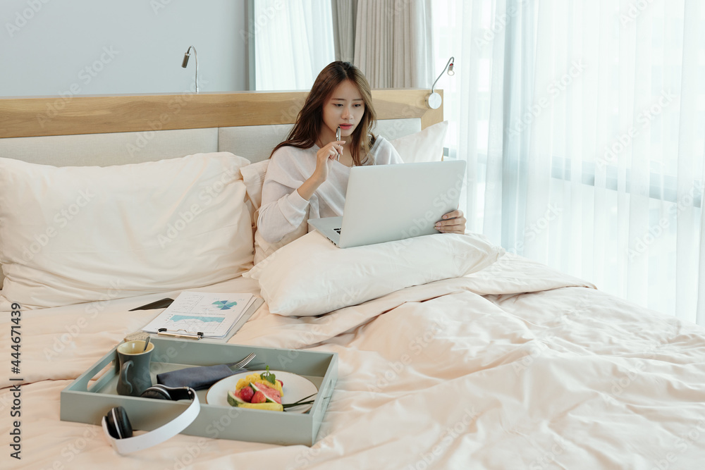 Pensive young manager of marketing department working from home due to coronavirus pandemic, she is sitting in bed and analyzing data on laptop screen Pensive young manager of marketing department working from home due to coronavirus pandemic, she is sitting in bed and analyzing data on laptop screen