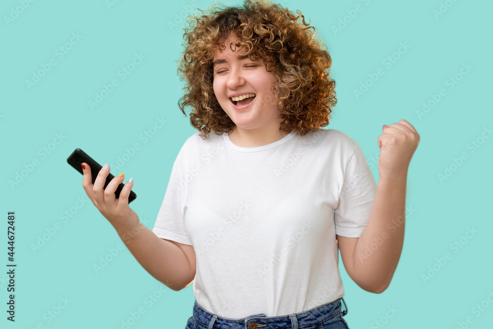 Happy phone news. Body positive. Diet app. Online communication. Excited emotional overweight young woman with curly hair received message isolated on blue background. Happy phone news. Body positive. Diet app. Online communication. Excited emotional overweight young woman with curly hair received message isolated on blue background.