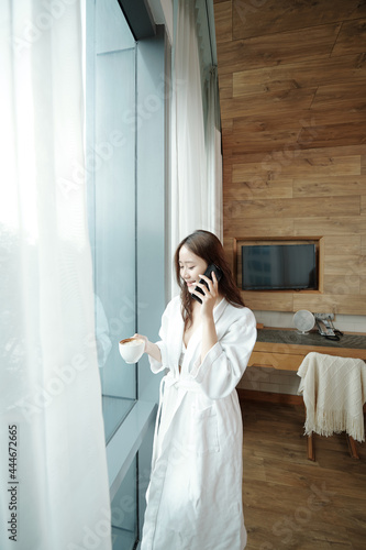 Happy pretty young woman in bathrobe standing at bedroom window, drinking coffee and talking on phone with boyfriend