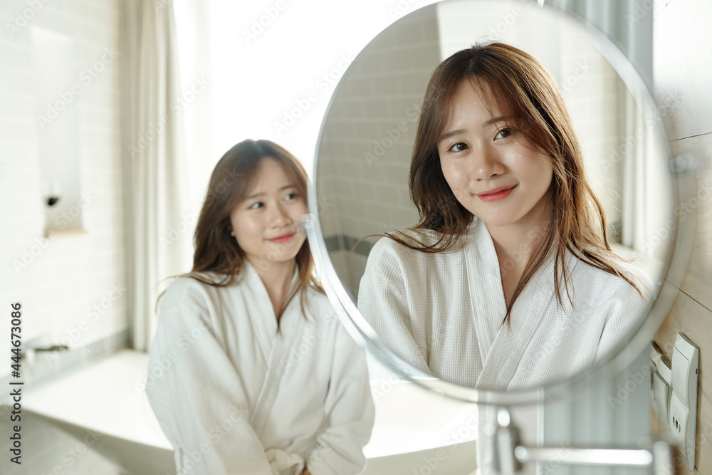 Fototapeta premium Smiling attractive young woman in bathrobe looking at magnifying mirror after taking shower
