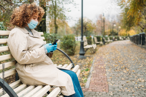 Corona communication. Covid leisure. Quarantine lifestyle. Curly hair overweight woman in medical face mask outside phone chatting in autumn park landscape copy space.
