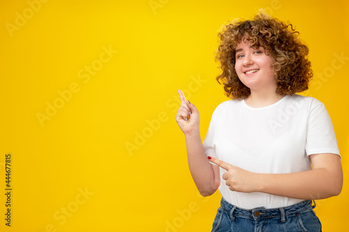 Pointing girl. Presenting product. Body positive. Advertising background. Happy smiling overweight woman with curly hair on yellow empty space.