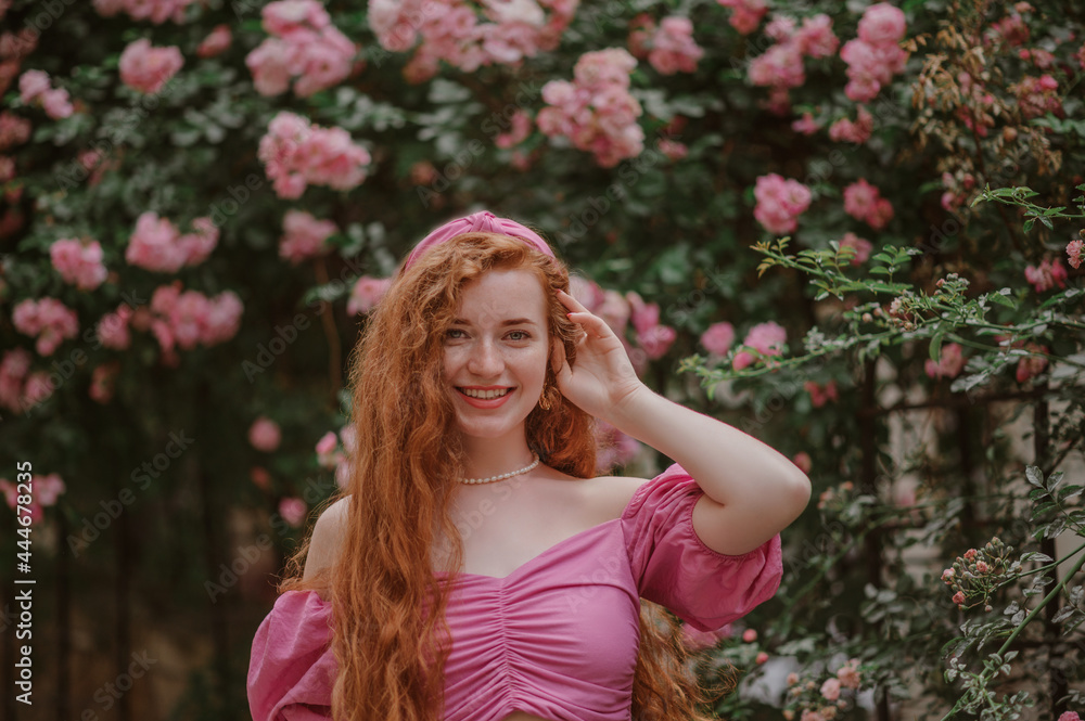 Beautiful happy smiling redhead woman with long natural curly hair wearing trendy summer pink blouse, headband, posing in blooming rose garden. Copy, empty space for text Beautiful happy smiling redhead woman with long natural curly hair wearing trendy summer pink blouse, headband, posing in blooming rose garden. Copy, empty space for text