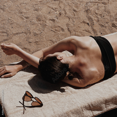 Beautiful young woman sunbathes on the beach. Female sunglasses on beige towel. Minimal aesthetic lifestyle fashion blog, magazine, social media. Sunbathe, relax, chill on summer travel vacations photo