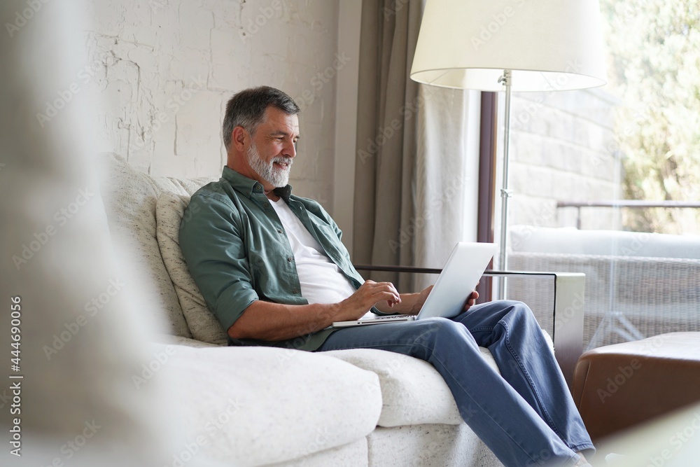 Portrait of happy mature man in casual clothes using laptop lying on sofa in house.