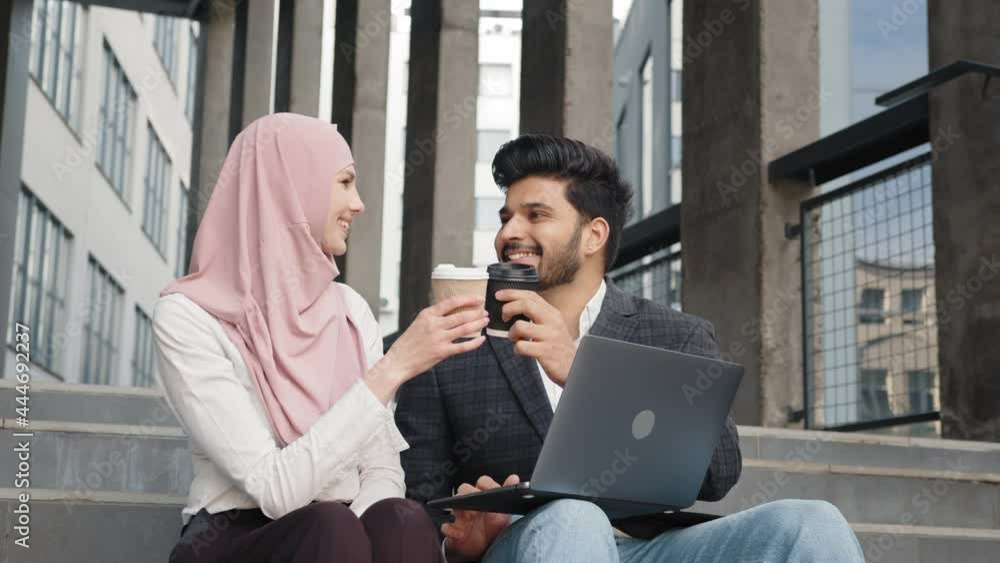 Happy muslim man and woman toasting with cups of coffee while sitting together on outdoor stairs. Business partners using laptop for meeting on fresh air. Muslim man and woman work on stairs Happy muslim man and woman toasting with cups of coffee while sitting together on outdoor stairs. Business partners using laptop for meeting on fresh air. Muslim man and woman work on stairs