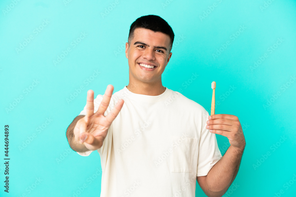 Young handsome man brushing teeth over isolated blue background happy and counting three with fingers Young handsome man brushing teeth over isolated blue background happy and counting three with fingers