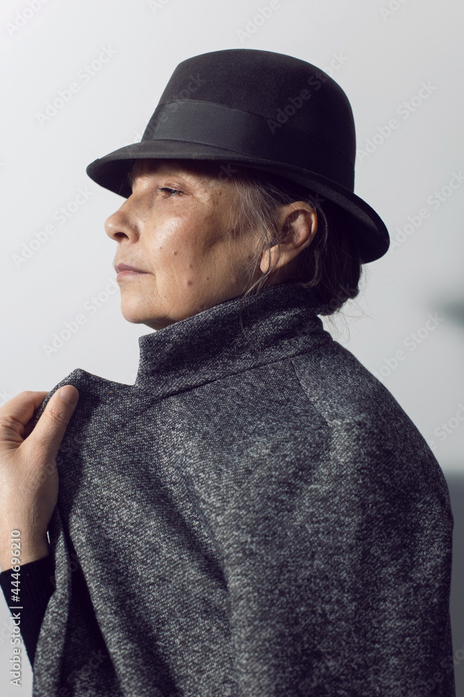 fashionable older woman in black clothes and a hat stands against a white wall in the office fashionable older woman in black clothes and a hat stands against a white wall in the office