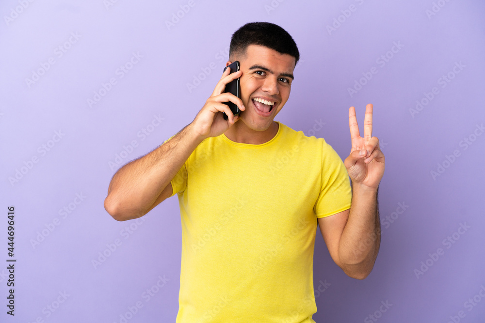 Young handsome man using mobile phone over isolated purple background smiling and showing victory sign Young handsome man using mobile phone over isolated purple background smiling and showing victory sign