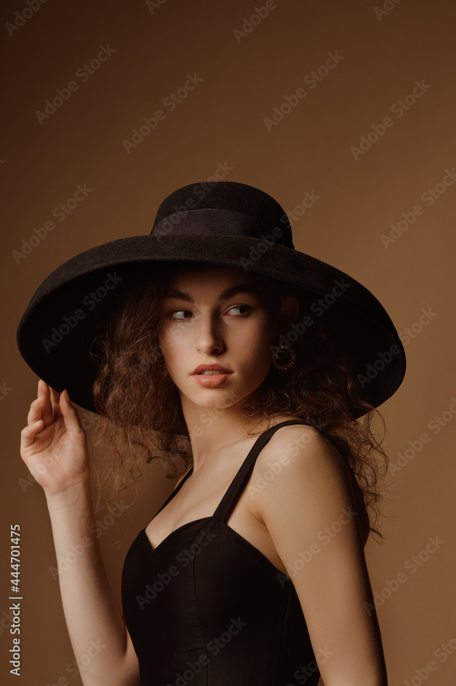 Fashion portrait of elegant curly brunette woman wearing luxury wide brim hat, black dress. Model looking aside Fashion portrait of elegant curly brunette woman wearing luxury wide brim hat, black dress. Model looking aside