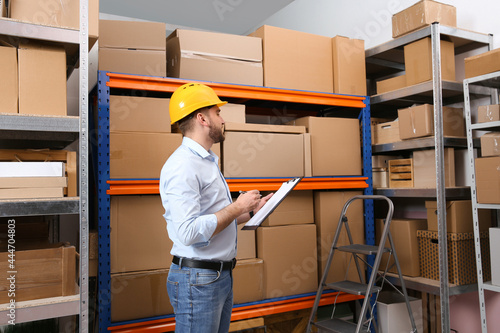 Young man with clipboard near rack of cardboard boxes at warehouse