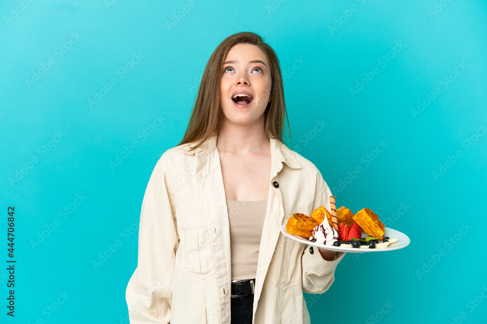 Teenager girl holding waffles over isolated blue background looking up and with surprised expression Teenager girl holding waffles over isolated blue background looking up and with surprised expression