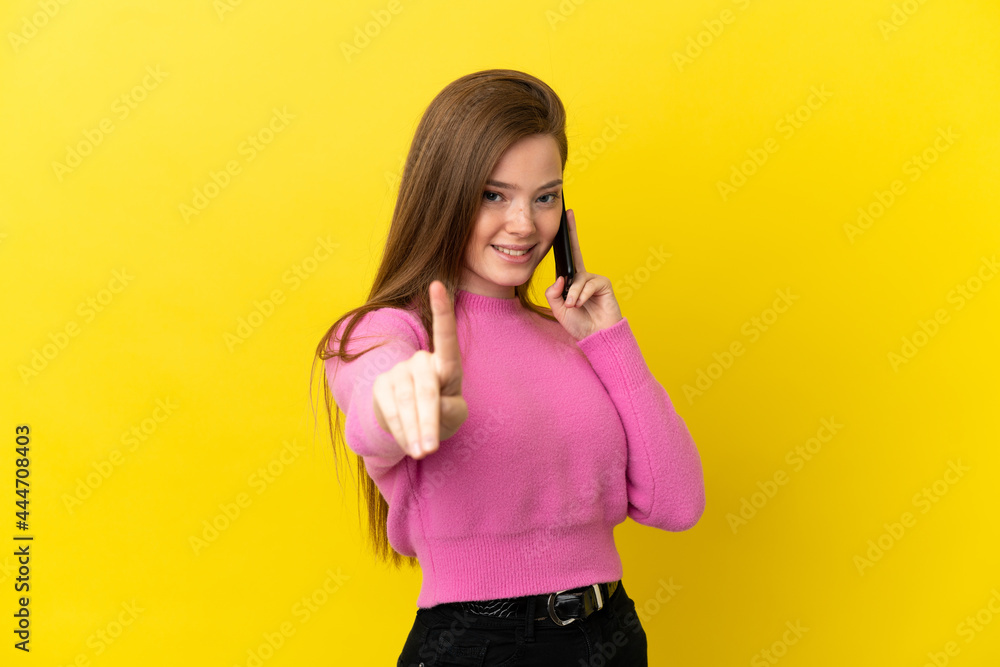 Teenager girl using mobile phone over isolated yellow background showing and lifting a finger Teenager girl using mobile phone over isolated yellow background showing and lifting a finger