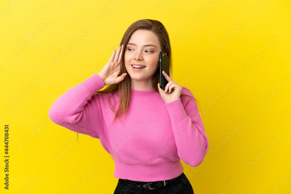 Teenager girl using mobile phone over isolated yellow background listening to something by putting hand on the ear Teenager girl using mobile phone over isolated yellow background listening to something by putting hand on the ear
