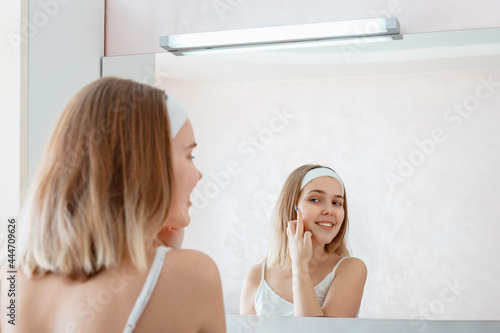 Young smiling woman apply skincare moisturizer body cream on face in bathroom after shower. Happy teenage girl do Self Care morning routine. Beauty spa treatment at home.