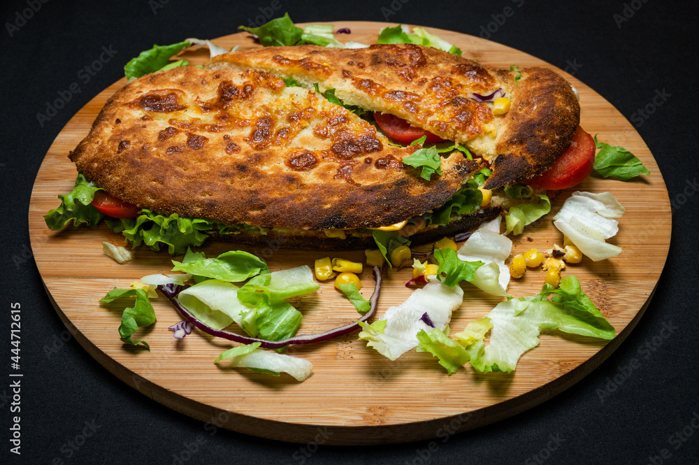 sandwich with mozzarella cheese, cedar cheese, mushrooms, red peppers, corn and iceberg lettuce on a wooden plate on a black background.