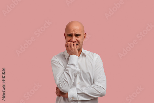 Can i tomorrow. Horizontal portrait of nervous bald man with bristle, bites finger nails anxiously, worried of go to the boss for an audience. Dressed in white shirt. Isolated on pink background.