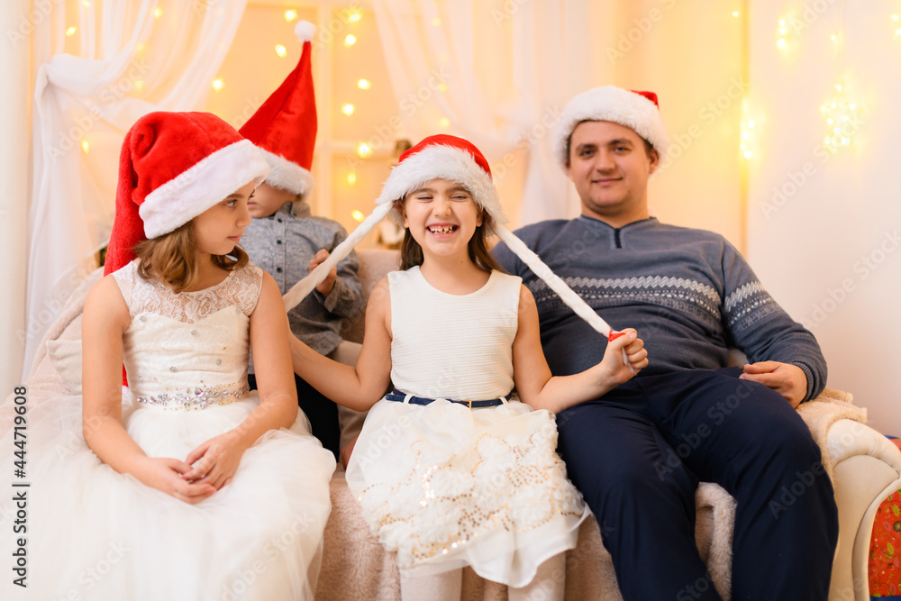 Fototapeta premium family portrait - parent and children in home interior decorated with holiday lights and gifts, dressed in santa hat for new year celebration