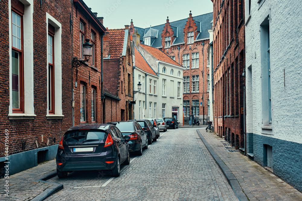 Brugge street with cobblestone road with parked cars and old medieval houses. Bruges, Belgium