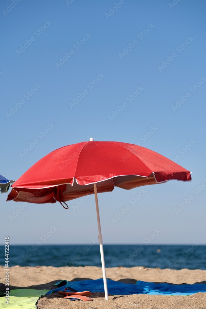 Red beach umbrella with blue towel and slippers on the sand with the sea and blue sky in the background