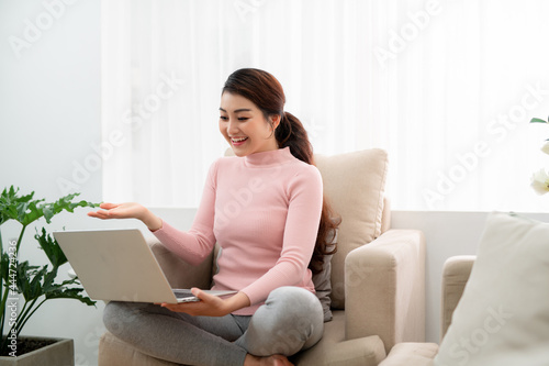 Beautiful cheerful girl using silver laptop while sitting on sofa in living room at home.