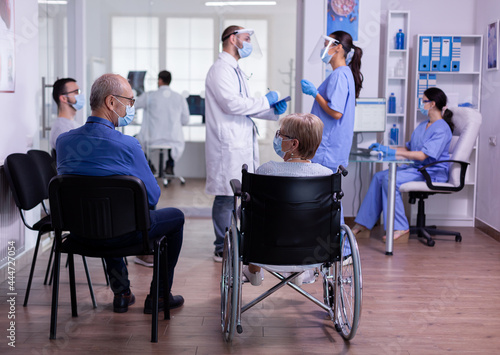 Specialist doctor with visor and protection mask speaking with assistant standing in waiting room of medical clinic before patients examination. People visiting medic during covid 19 global pandemic