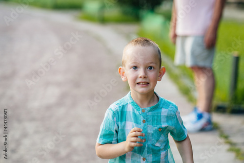 Portrait of blond excited little blond caucasian boy four years old looking at camera while standing path in the summer park, her grandfather on blurred background.