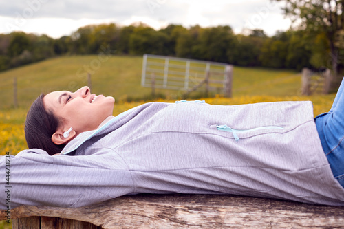 Woman Lying On Bench In Countryside Relaxing And Listening To Music Or Podcast On Wireless Earphones