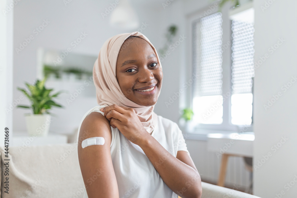 Portrait of a muslim African female smiling after getting a vaccine. Woman holding down her shirt sleeve and showing her arm with bandage after receiving vaccination. Portrait of a muslim African female smiling after getting a vaccine. Woman holding down her shirt sleeve and showing her arm with bandage after receiving vaccination.