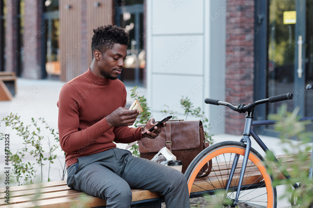 Serious African white collar worker eating sandwich and using smartphone Serious African white collar worker eating sandwich and using smartphone