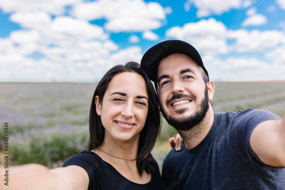 Fototapeta premium Smiling couple hugging each other taking a selfie with smart phone on lavender field - Joyful young tourists having fun and posing on camera self portrait while standing outdoors