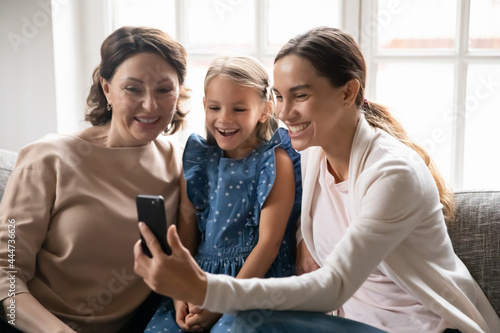 Smiling little girl with young mother and old grandmother talk speak on video call cellphone relax at home. Happy three generations of women use smartphone have webcam digital virtual event.