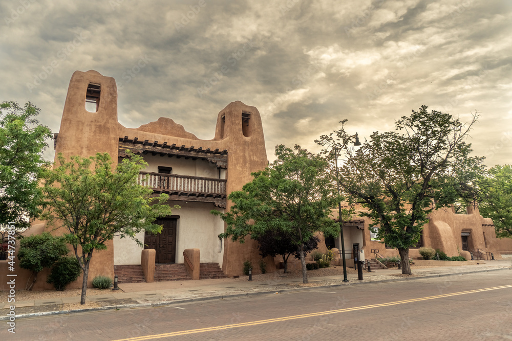 Pueblo Revival Style building with earth tone color, rounded corners and battered walls under dramatic cloudy sky, front, New Mexico Museum of Art, Santa Fe, New Mexico