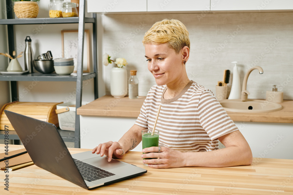 Young businesswoman with vegetable smoothie looking at screen of laptop Young businesswoman with vegetable smoothie looking at screen of laptop