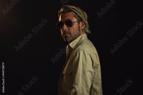 Man with blond hair and a stubble beard and dark aviator sunglasses wears a safari shirt. Looking over his shoulder.