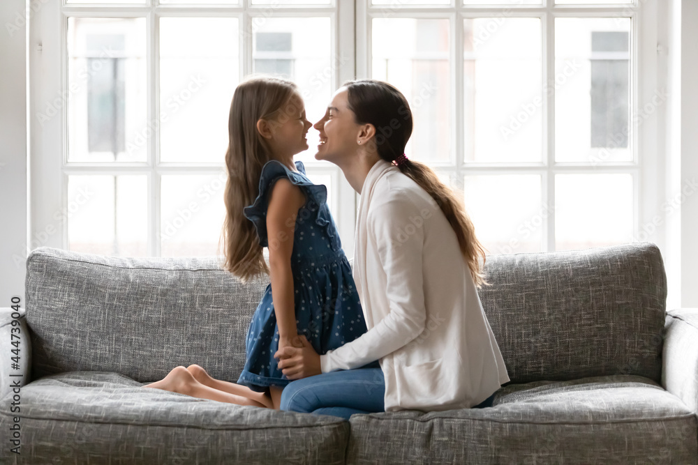 Loving young Caucasian mother and little teen daughter sit relax on sofa in living room play on family weekend. Happy mom and small girl child rest at home feel playful enjoy close moments together.
